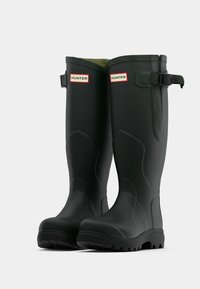 Juodi guminiai lietaus batai su lygia tekstūra, su "HUNTER" logotipu ir reguliuojamu dirželio detalumu ant viršaus. Tvirta guminė išorinė pusė su raštu.