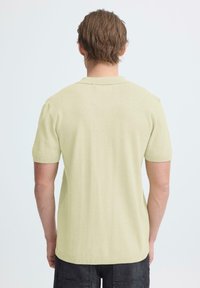 Man met kort bruin haar die een lichtgeel poloshirt met korte mouwen en een donkere broek draagt, staand met zijn rug naar de camera tegen een effen achtergrond.