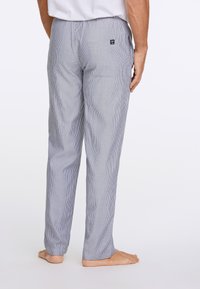 Man in een lichtgrijze streepjesshort en een wit shirt, staand op blote voeten tegen een plain witte achtergrond.