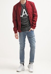 Rote Bomberjacke mit gerippten Bündchen, Reißverschlusstaschen, schwarzes Grafik-T-Shirt darunter, schlanke hellblaue Jeans und weiße Sneakers.