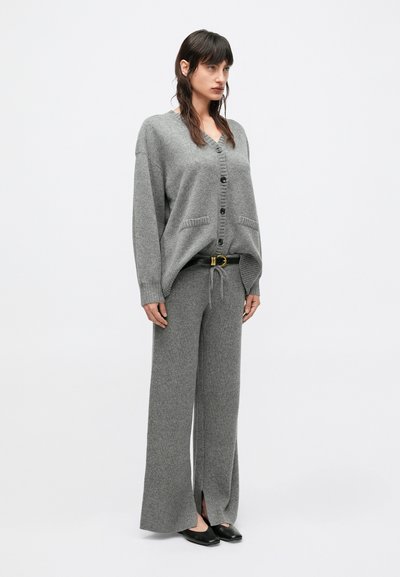 Szary kardigan z guzikami, połączony z szarymi szerokimi spodniami z wiązaniem. Stylizację dopełnia czarny pasek i płaskie buty.
