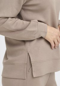 Sweatshirt beige avec une coupe décontractée, un tissu doux, des fentes sur les côtés à l'ourlet et des accents cousus sur les poignets et le bas.
