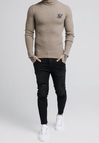 Beige gerippter Rollkragenpullover mit kleinem schwarzem Logo, kombiniert mit schwarzen, zerrissenen Skinny Jeans und weißen Turnschuhen.