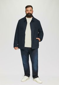 Dunkelblauer Blazer mit sichtbaren Knöpfen und Seitentaschen, getragen über einem hellen Pullover. Kombiniert mit dunklen Jeans und weißen Sneakers.