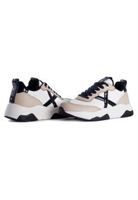 Beige and white sneakers feature a textured finish, black accents, a thick sole, and black laces, with a branded heel tab.