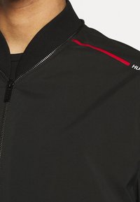 Giacca bomber nera con colletto a coste, chiusura con zip e una linea di accento rossa sulla spalla; texture liscia e materiale leggero.