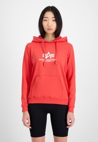 Sudadera color rojo coral con bolsillo frontal, puños acanalados y capucha con cordón; presenta un logo blanco en el pecho. Combinada con shorts negros.
