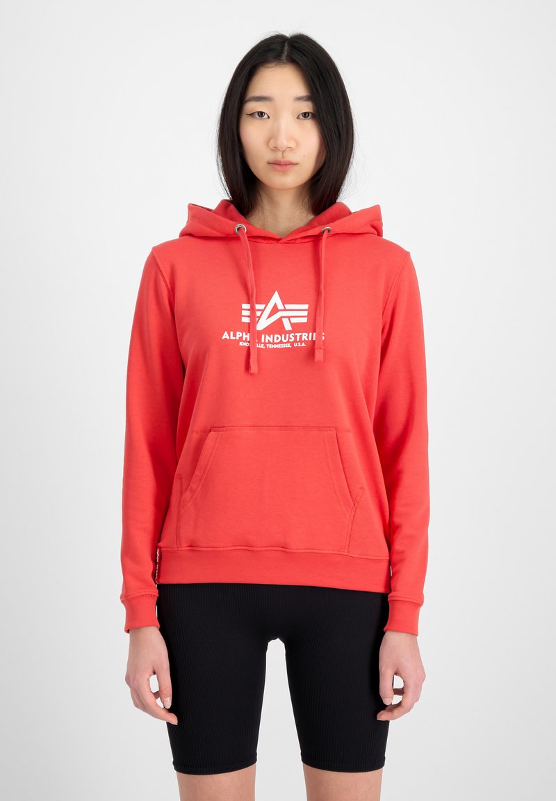 Sudadera color rojo coral con bolsillo frontal, puños acanalados y capucha con cordón; presenta un logo blanco en el pecho. Combinada con shorts negros.