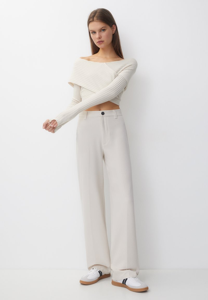 Giovane donna che indossa un maglione crema a coste con spalle scoperte, pantaloni crema a vita alta e gamba larga, e sneaker bianche con suola marrone.