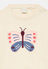 Cream-colored shirt with a large embroidered butterfly design. The butterfly features pink and blue wings with dark accents and detailing.