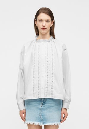 Kobieta ubrana w białą bluzkę z długim rękawem z koronkowym zdobieniem i falbankowanym kołnierzem, w połączeniu z jasnoniebieską, postrzępioną dżinsową spódnicą, stojąca prosto.