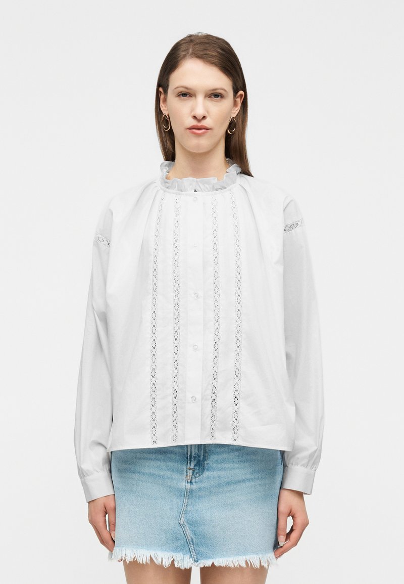 Γυναίκα που φοράει άσπρη μπλούζα με μακριά μανίκια, λεπτομέρειες από δαντέλα και γιακαδάκι με βολάν, συνδυασμένη με φούστα τζιν ανοιχτού μπλε χρώματος με ξεφτισμένα άκρα, στέκεται όρθια.