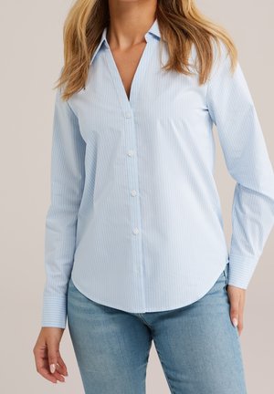 Femme portant une chemise à rayures fines bleu clair avec des boutons et un jean bleu, debout, les mains détendues le long du corps.