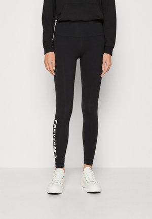 Leggings negras con una cinturilla alta que presentan el logo de "Converse" impreso en blanco a lo largo de la pierna izquierda. Combinados con zapatillas blancas.