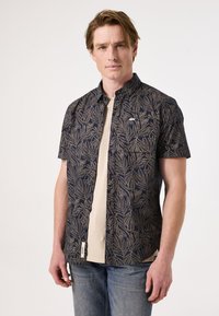 Korte mouw shirt in marineblauw met een bladenpatroon, gemaakt van lichtgewicht stof. Voorzien van een knoopsluiting aan de voorkant en een borstzak. Gedorven over een beige T-shirt.