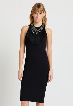 Jeune femme aux cheveux blonds portant une robe midi noire sans manches et un collier noir à franges avec des perles, debout devant un fond blanc.