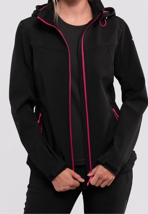 Vrouw draagt een zwart jack met roze rits en accenten op de zakken, houdt de ritssluiting bij de taille vast tegen een neutrale achtergrond.