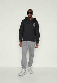 Sweatshirt preta com capuz, bolso frontal e logotipo, combinado com calças de fato cinzas. O modelo usa óculos de sol e ténis cinzentos claros.