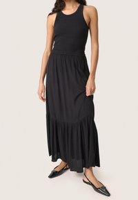 Débardeur noir côtelé avec une longue jupe noire fluide présentant un ourlet volé. Le modèle porte des ballerines noires brillantes à bout pointu.