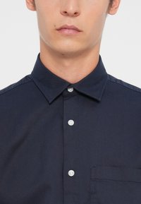 Tumši zilas krāsas krekls ar apkakli, ar pogām, baltām pogām un krūšu kabatu; izgatavots no gludas, audētas auduma.