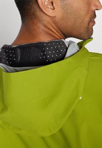 Zielona wodoodporna kurtka z teksturowanym materiałem, posiadająca czarny kołnierz wewnętrzny w białe kropki i wypukłe logo na plecach.