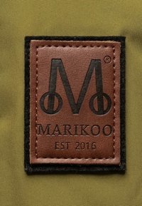 Smeđa kožna oznaka s crnim šavovima koja prikazuje slovo "M" i "MARIKOO EST 2016", smještena na maslinastozelenoj tkanini.
