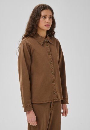 Jeune femme aux longs cheveux bouclés portant une chemise marron boutonnée et un pantalon marron assorti sur un fond clair uni.