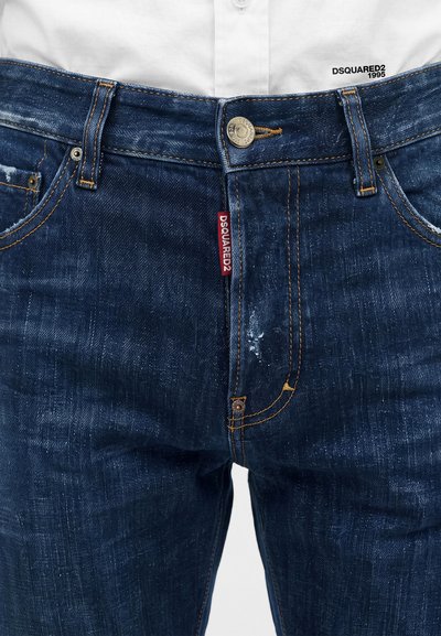Gros plan avant d'un jean en denim bleu porté avec une chemise blanche, montrant l'étiquette DSQUARED2 et le logo sur l'ourlet de la chemise.