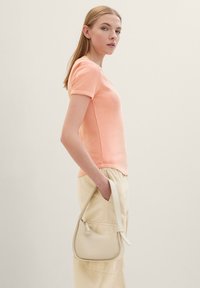 Perzikkleurig geribbeld T-shirt met korte mouwen, gecombineerd met beige hoge taillebroeken; een kleine crèmekleurige handtas met een ritsdetail rust aan de zijkant.