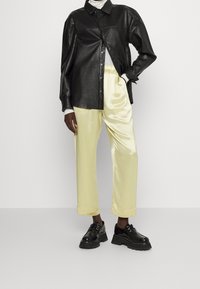Svart läder skjorta med knappar, ljusgul satinbyxor och kraftiga svarta skor. Slät textur och kontrasterande färger med en avslappnad passform.