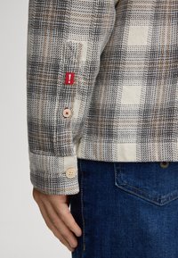 Camicia di flanella a quadri con tonalità di grigio, crema e marrone. Presenta due bottoni sui polsi e un'etichetta rossa come dettaglio. Texture morbida.