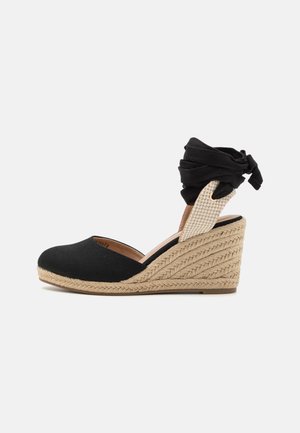 Sandales compensées en tissu noir avec une semelle en jute tissée et une lanière à la cheville, dotées d'un bout fermé et d'une sangle élastique à motifs.