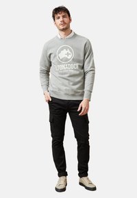 Sudadera gris con un logo, cuello redondo y puños acanalados, combinada con pantalones cargo negros y zapatillas beige.