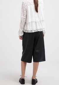 Blouse blanche à volants avec détails en dentelle, associée à un pantalon culotte en simili cuir noir. Le modèle porte des chaussures noires, ses cheveux tombent au-dessous des épaules.