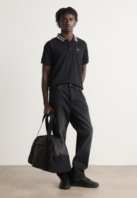Camisa polo negra con cuello de rayas blancas, combinada con jeans de mezclilla oscuros. La persona lleva una bolsa de lona en tonos negro y gris.
