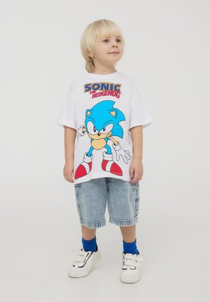 T-shirt bianca con una grande grafica blu di Sonic the Hedgehog, abbinata a pantaloni shorts in denim chiaro e sneakers bianche con dettagli blu e neri.