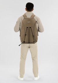 Mochila marrón fabricada con material duradero, que cuenta con correas ajustables, un asa en la parte superior y un cierre de cordón en la parte frontal.