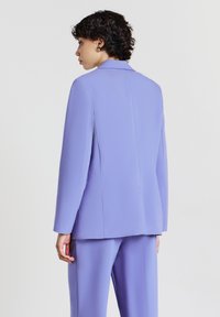 Persona con i capelli ricci corti, che indossa un giacca su misura lavanda e pantaloni coordinati, di spalle contro uno sfondo neutro.