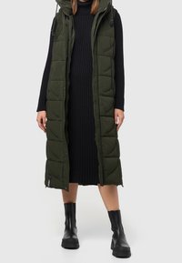 Długa, bezrękawnikowa, oliwkowozielona pikowana kamizelka z przeszywanymi sekcjami, z przednim zamkiem błyskawicznym i kapturem, noszona na czarnej sukience w prążki oraz czarnych masywnych butach.