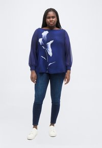 Γυναίκα με μακριά κοτσίδια, ντυμένη με διάφανη μπλε μπλούζα με λευκό λουλουδάτο σχέδιο, σκούρο μπλε τζιν και λευκά αθλητικά παπούτσια, που στέκεται σε απλό φόντο.