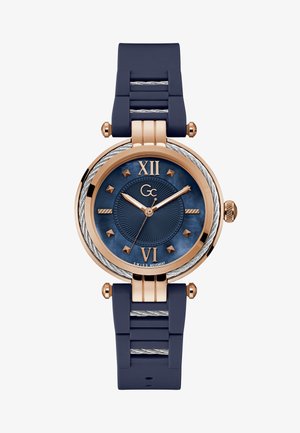 Kerek óra tengerészkék gumiszíjjal és rózsa arany színű akcentussal. Jellemzői között szerepel egy texturált kék számlap, római számokkal 12 és 6 óránál, valamint ezüst színű kiegészítők.