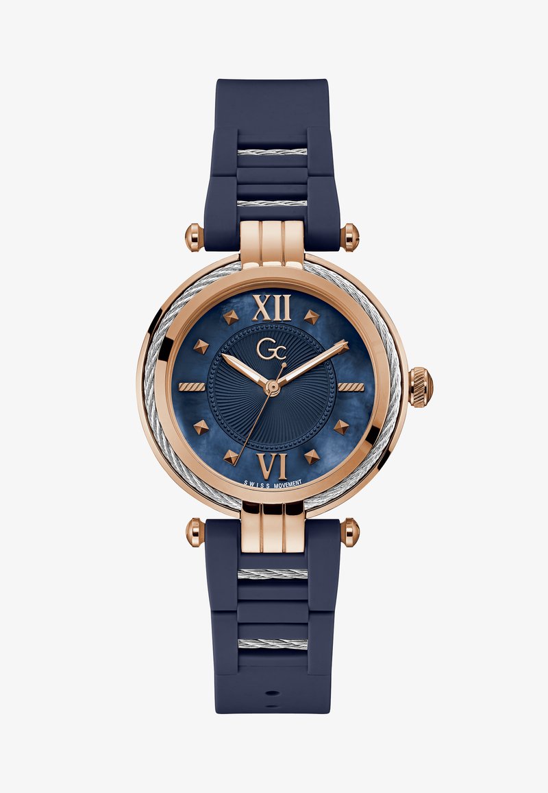 Ümmargune kell, millel on tumesinine kummirihm ja roosa kuldne viimistlus. Omab tekstureeritud sinist numbrit, Rooma numbreid kell 12 ja 6 ning hõbedaseid detaile.