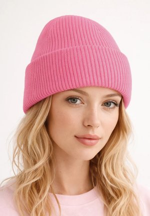 Young woman with blonde wavy hair wearing a bright pink ribbed knit beanie and a light pink top, neutral expression, white background.