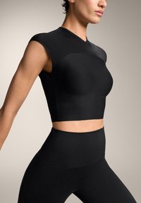 Črn športni top z kratkimi rokavi, z v-izrezom in gladko teksturo. Kombiniran z visokim pasom črnih pajkic.