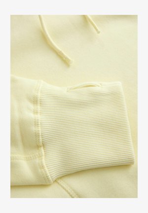 Kollane pusa kangas soonikutega mansettidega, näidates sujuvat tekstuuri ja nööridega. Omadused on pehme, kergkaaluline materjal mugavuse tagamiseks.