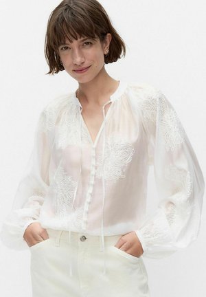 Femme aux cheveux courts bruns portant un chemisier blanc transparent avec broderie florale et un pantalon blanc, les mains dans les poches, souriant légèrement.
