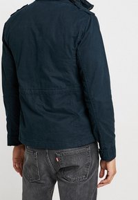 Mörkblå bomullskavaj med krage och axelremmar, med rak skärning och synliga sömdetaljer, i kombination med grå jeans.