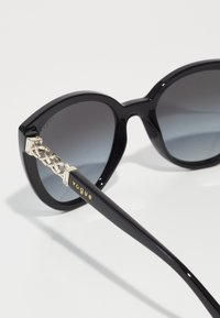 Ochelari de soare negri cu lentile gradient, având un accent împletit din aur pe tâmplă și cu "vogue" embosat în aur pe braț.