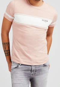 Camiseta de algodón de color rosa claro con una franja blanca horizontal y el logotipo negro "GUESS". Diseño de mangas cortas y cuello redondo. Combinada con jeans de color gris claro.