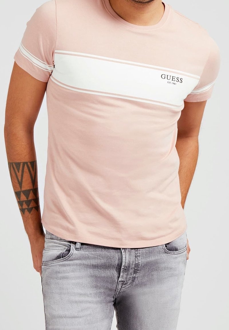 Camiseta de algodón de color rosa claro con una franja blanca horizontal y el logotipo negro "GUESS". Diseño de mangas cortas y cuello redondo. Combinada con jeans de color gris claro.